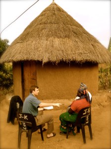 Two people sitting outside a traditional thatched-roof hut for discussion.