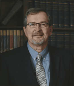 Man in glasses and suit in front of a bookshelf, speaking or teaching.
