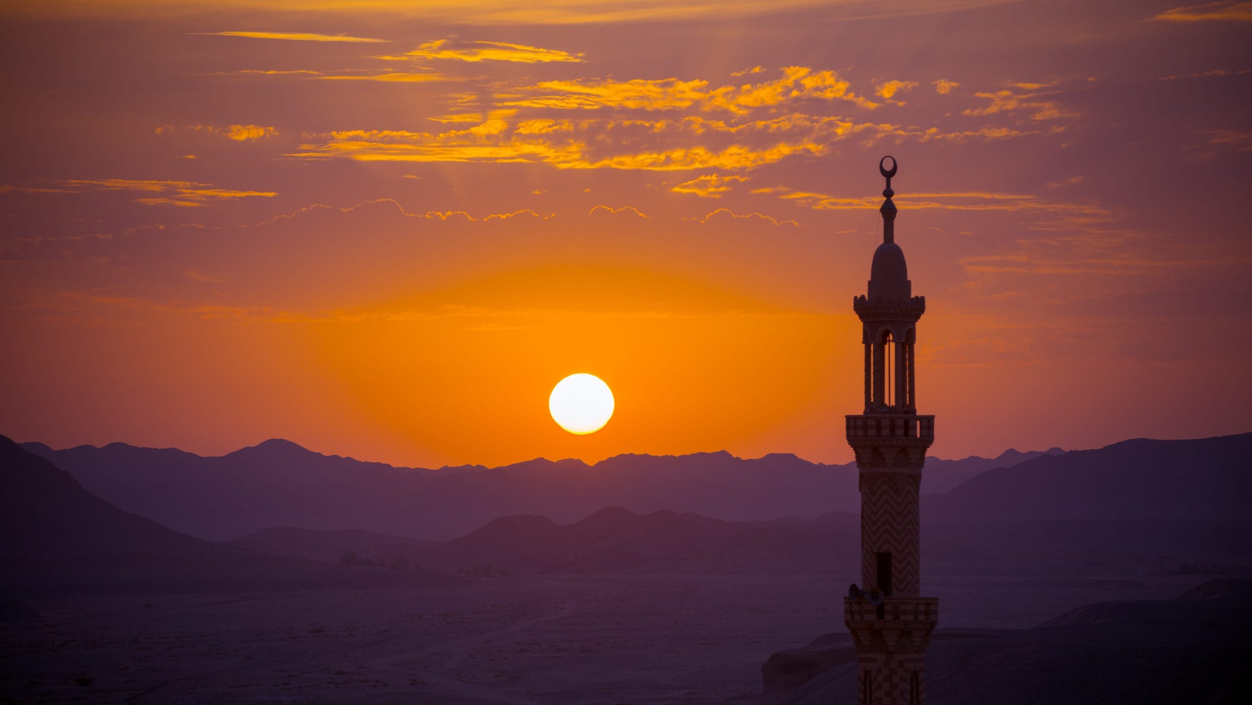 Sunset over desert landscape with a tall minaret silhouette.