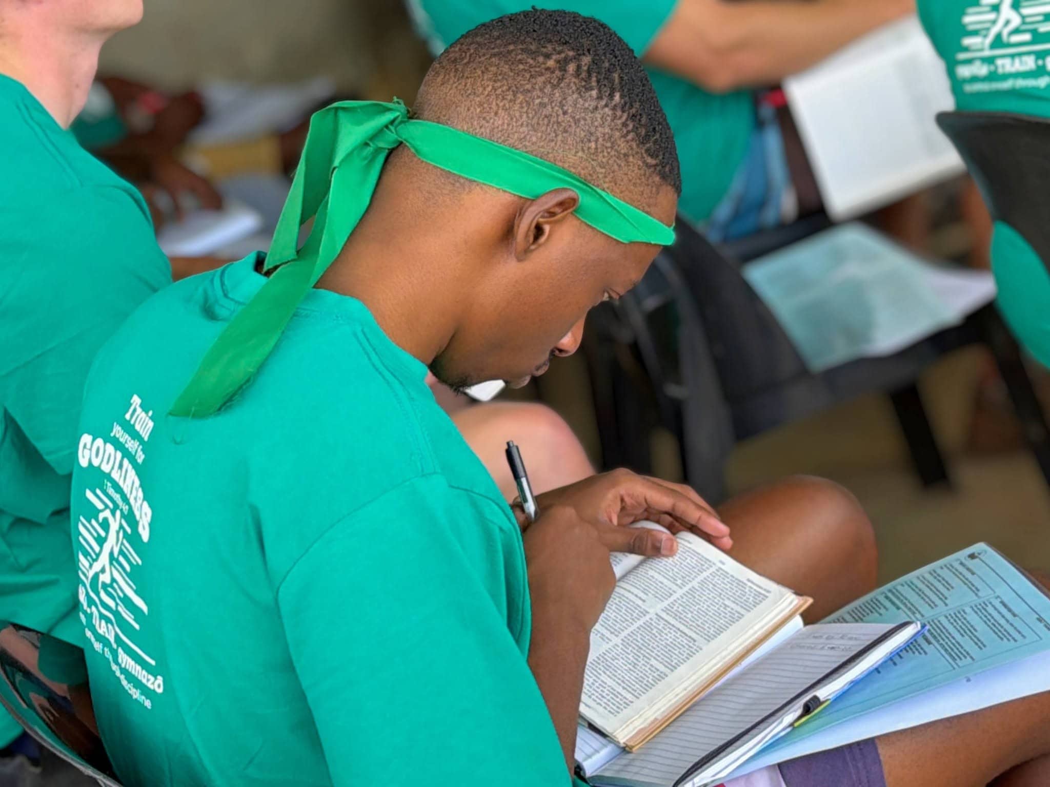 Young man in a green Godmakers shirt reading the Bible during a Christian camp study session.