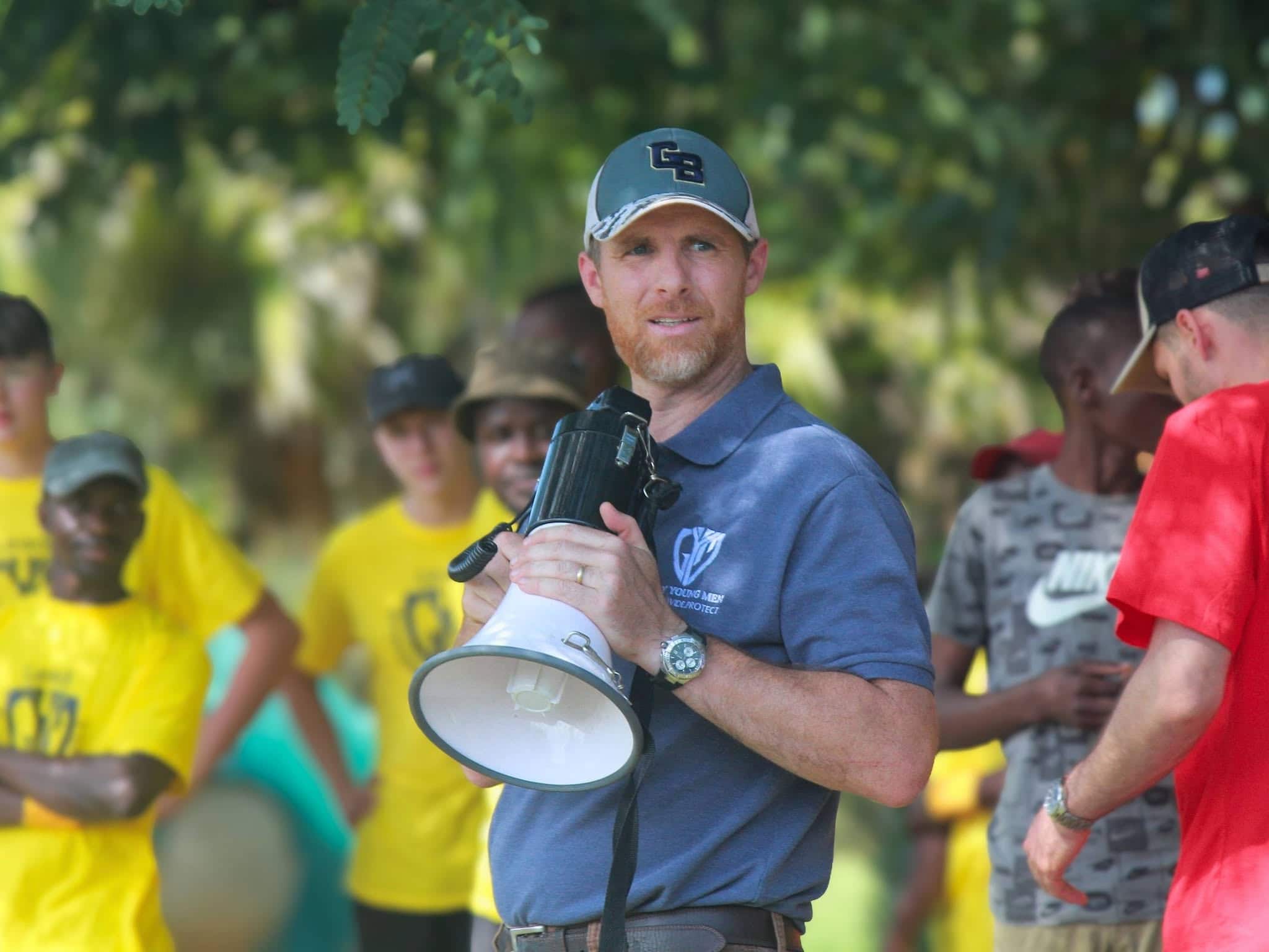 Christian youth camp leader holding a megaphone while speaking to young participants outdoors.
